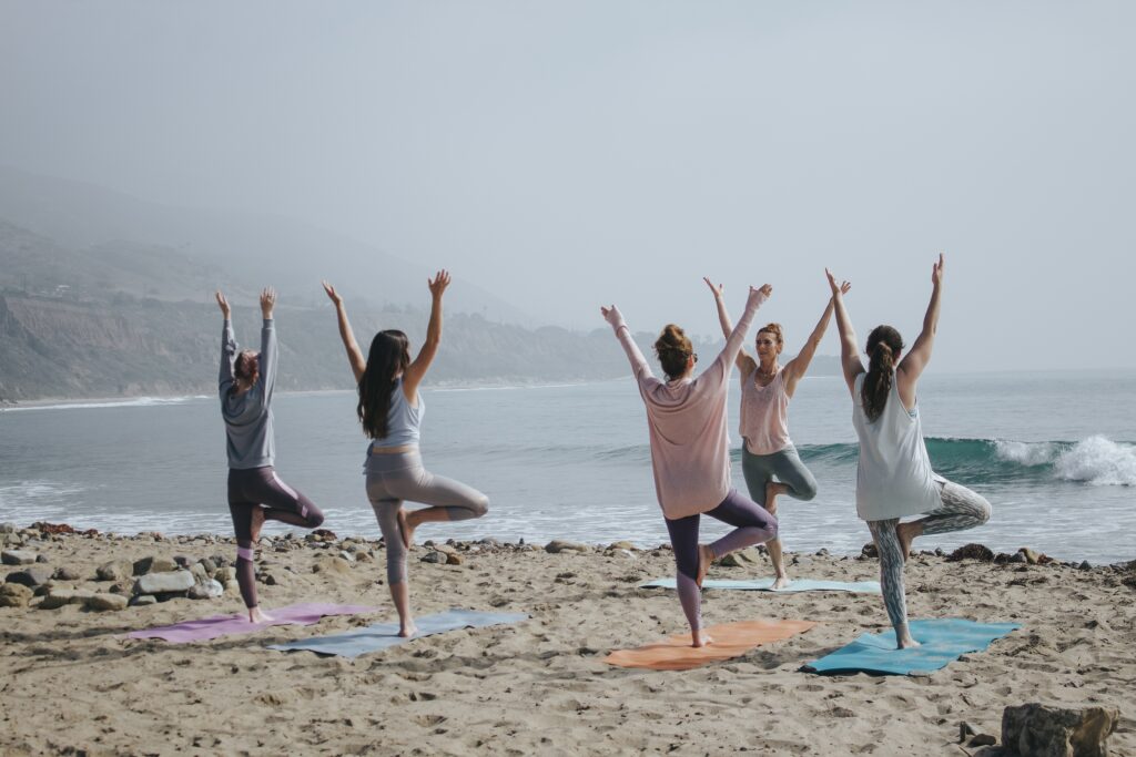 Corporate Wellness Retreat at a beach. Five women doing yoga on mats in the sand near the shore.