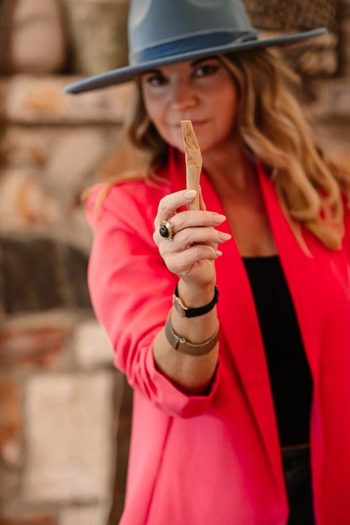 Amanda Rieger Green, wearing a pink blazer and grey hat. She's holding a piece of palo santo in her hand and smiling. She is practicing her energetic and spiritual health.